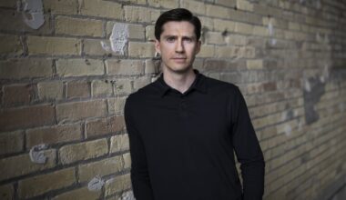 A man wearing a dark shirt is pictured against a brick wall. He is looking directly into the camera. with a serious facial expression.