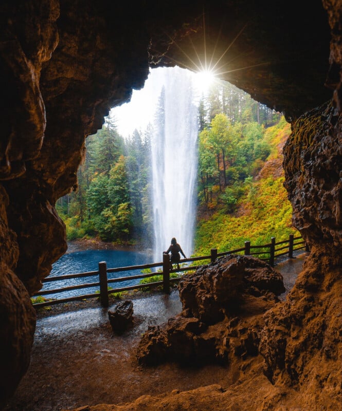A person stands by a wooden fence, viewing a tall waterfall through the mouth of a rocky cave, with green trees and sunlight filtering through the forest in the background.