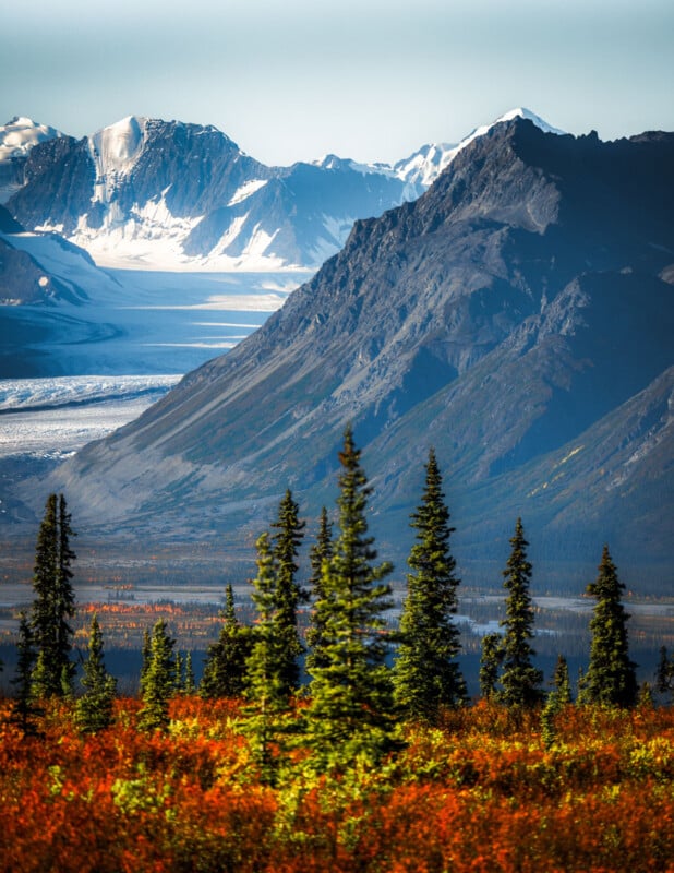 Snow-capped mountains rise behind dark rocky peaks, with a glacier visible in the valley. Evergreen trees and vibrant autumn foliage fill the foreground under a clear sky.