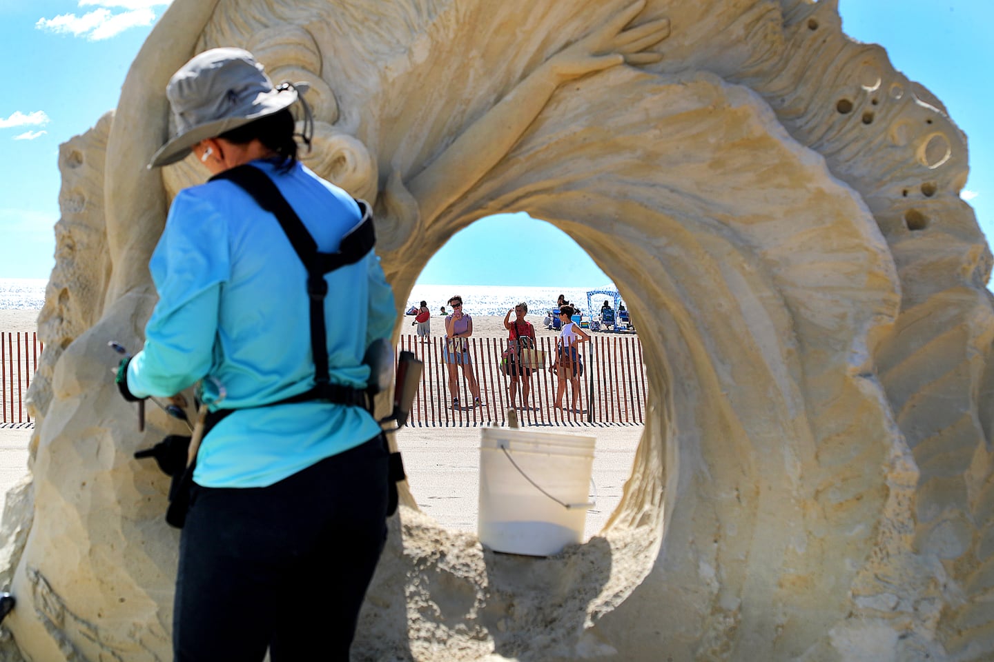 Onlookers stand on Hampton Beach as they are framed as Meliniege Beauregard from Captain Cook, Hawaii, works on her sand sculpture called "Go with the Flow."