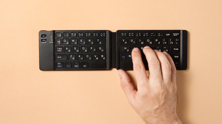 Hand typing on a compact foldable Bluetooth keyboard on a beige surface.