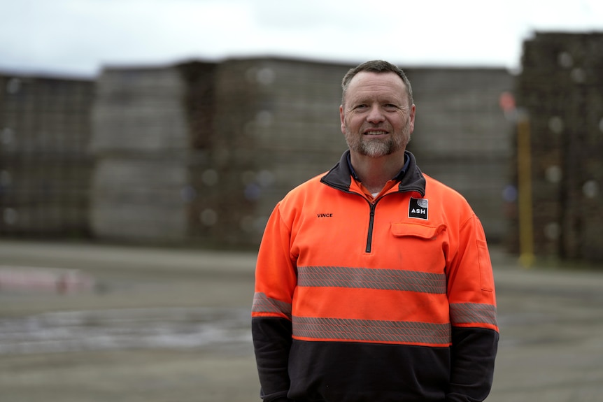 A man wearing a hi-vis orange jumper smiles as he stands outdoors. Behind him are stacks of timber.