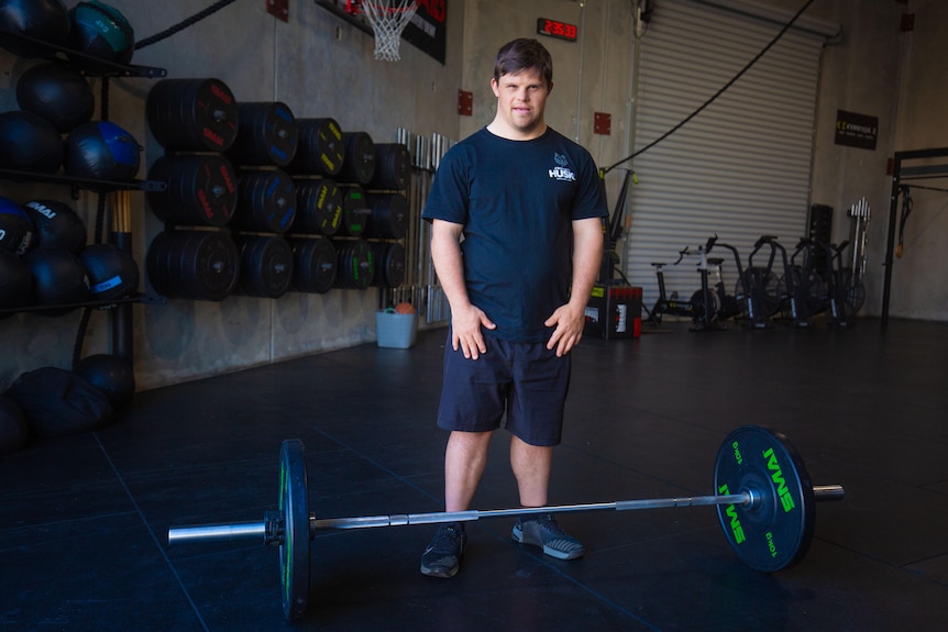 A young man with Down syndrome stands behind a loaded barbell inside a gym