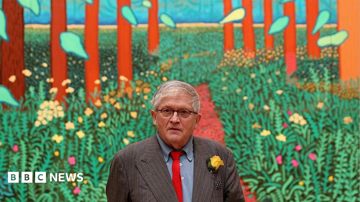 A man with grey hair and wearing black-rimmed glasses is standing in front of a colourful painting depicting green plants and tall trees. He is wearing a grey pin-striped suit, blue shirt and red tie with a yellow flower in his lapel.