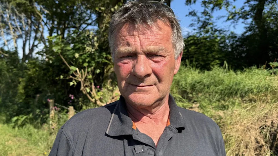Man looking troubled at the camera. He has short black and grey hair and is wearing a t-shirt. Behind him is green hedgerow 