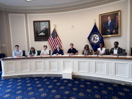 Students sit behind a curved committee-style desk in a government meeting room, participating in a discussion or briefing. American and state flags stand behind them, with framed portraits displayed on the walls.