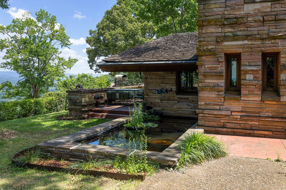 Stone house with a pond and garden, nestled among trees and greenery under a blue sky.