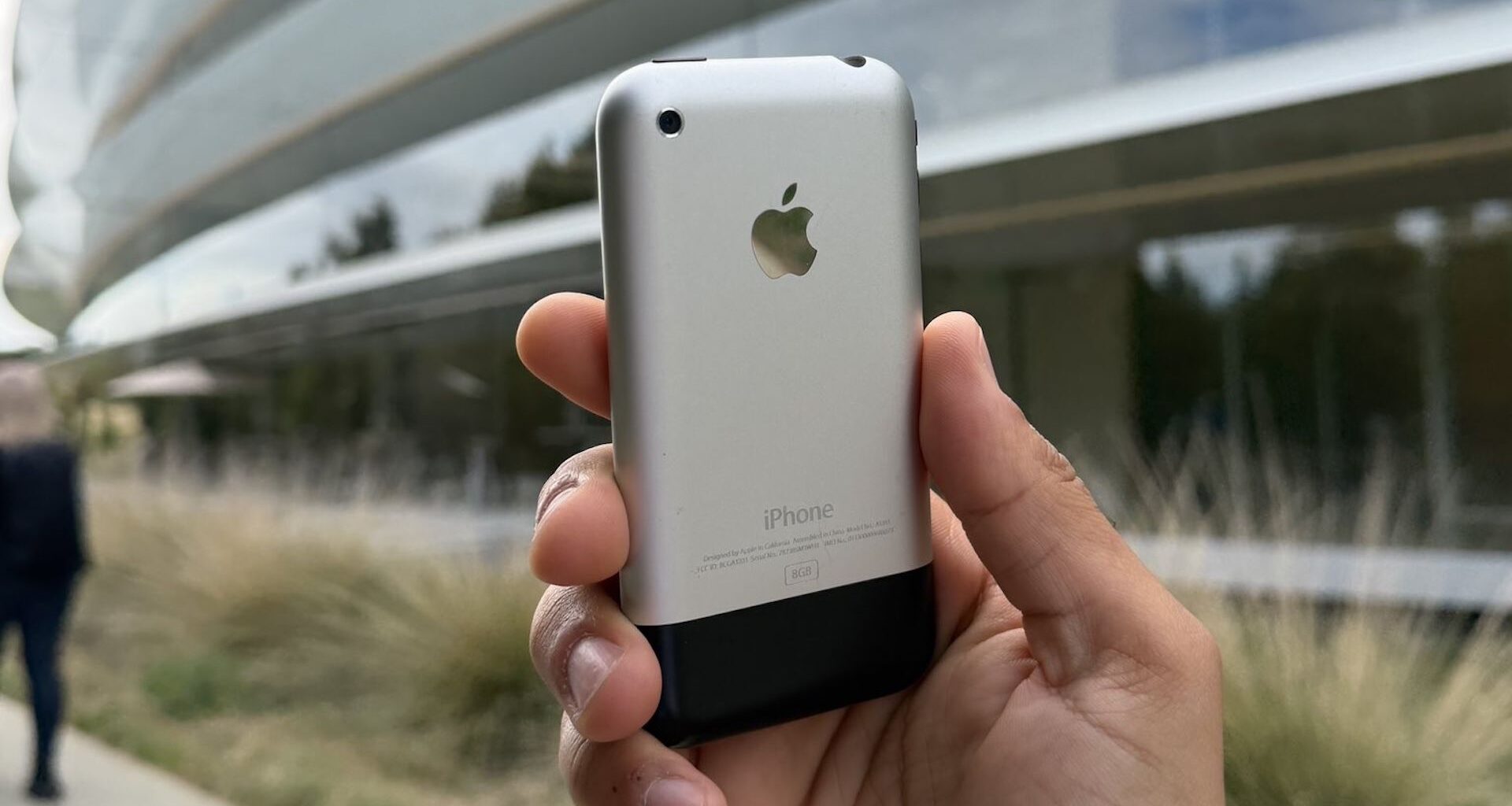 A hand holding up the original iPhone outside Apple Park