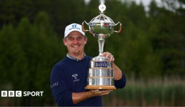 Bud Cauley, wearing a dark blue jumper and white cap, smiles and poses with the silver trophy to his left