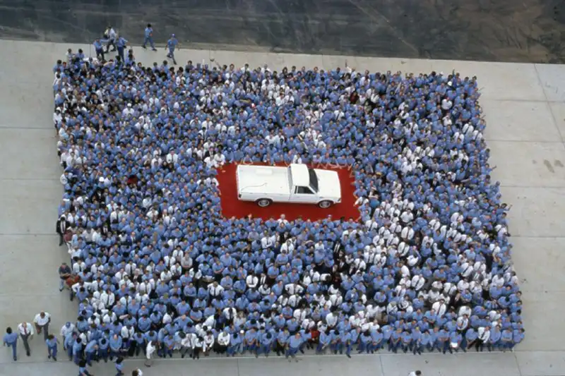 A white Nissan truck on a red platform with Nissan employees surround in a rectangle formation.