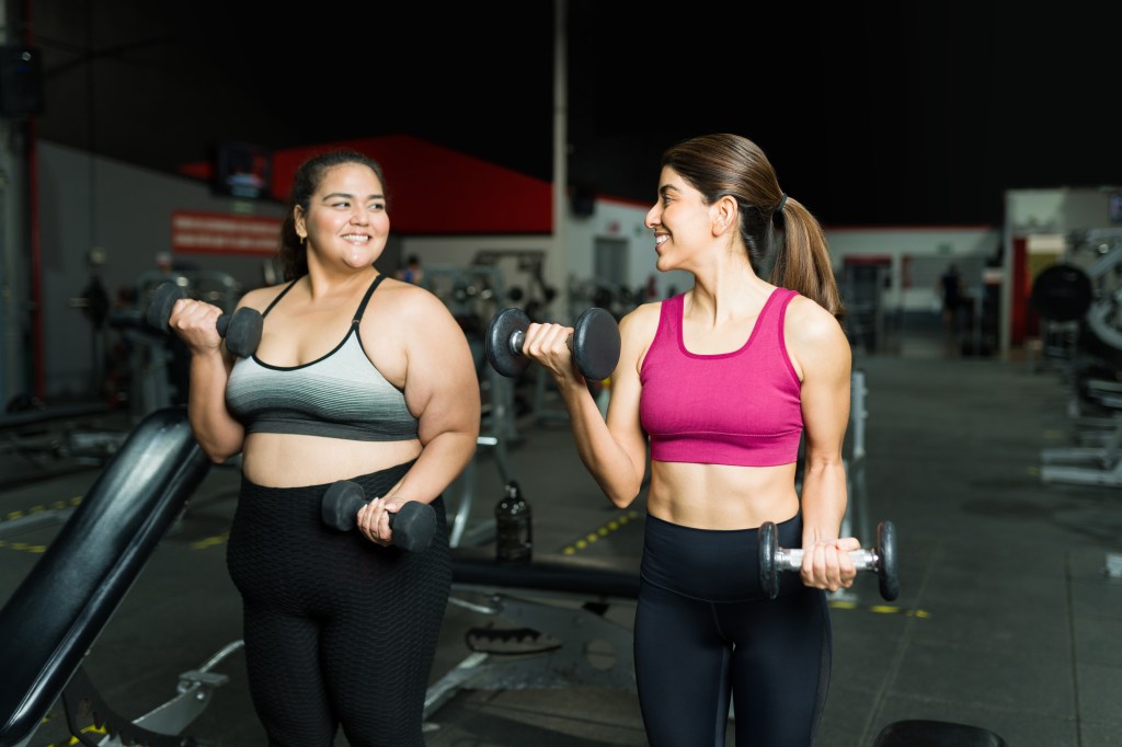 A smiling Hispanic personal trainer coaching an overweight woman lifting dumbbells in a gym.