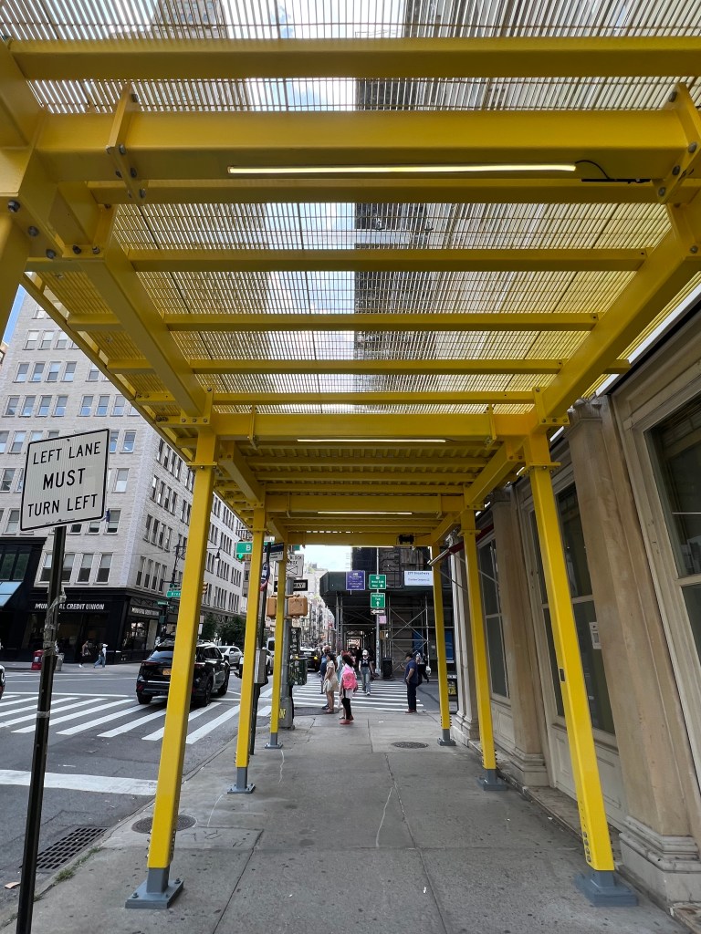 A new yellow sidewalk shed with an illuminated grated roof over a city sidewalk.