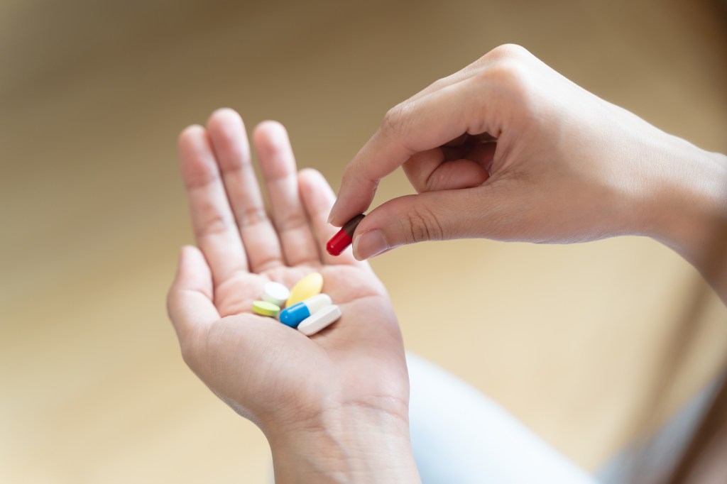 A person holding various pills and capsules in one hand, picking up a red and brown capsule with the other hand.