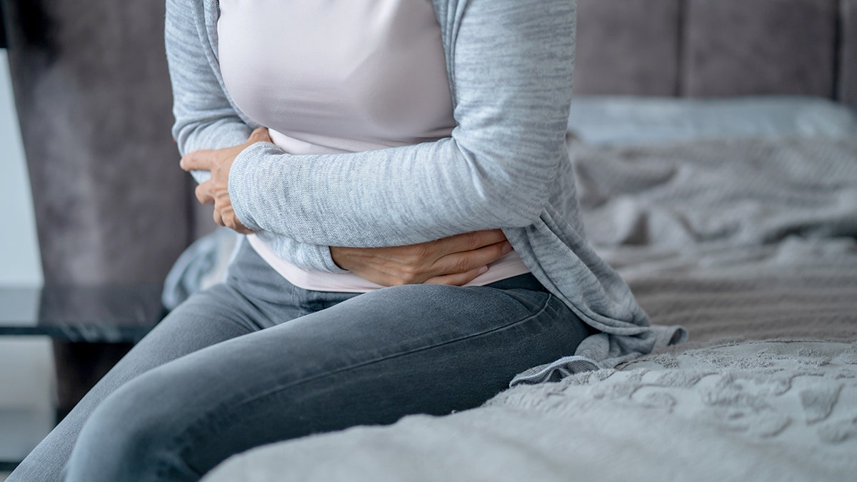 Woman holding her stomach and sitting on a bed appearing to experience abdominal pain.