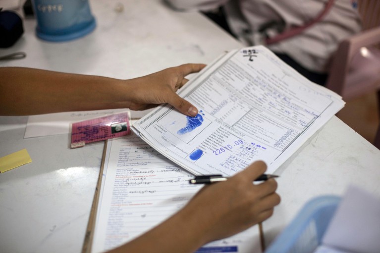 Hands holding and filling out official birth registration documents on a desk, with visible blue ink stamps and a pen at the Mae Tao Clinic in Thailand.
