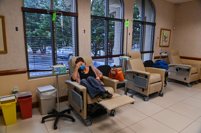 A woman wearing a face mask sits curled up with her head in her hand in a reclining medical chair in a clinic room, connected to an IV drip on a stand. Additional empty treatment chairs line the wall beside large windows next to her, with medical equipment and disposal bins visible around the room