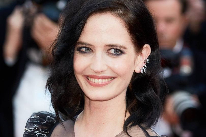 An individual with short, dark hair and a radiant smile, dressed in a sophisticated outfit, is standing amidst a crowd, seemingly engaging in conversation with the camera. The background is blurred, indicating a bustling event or gathering.