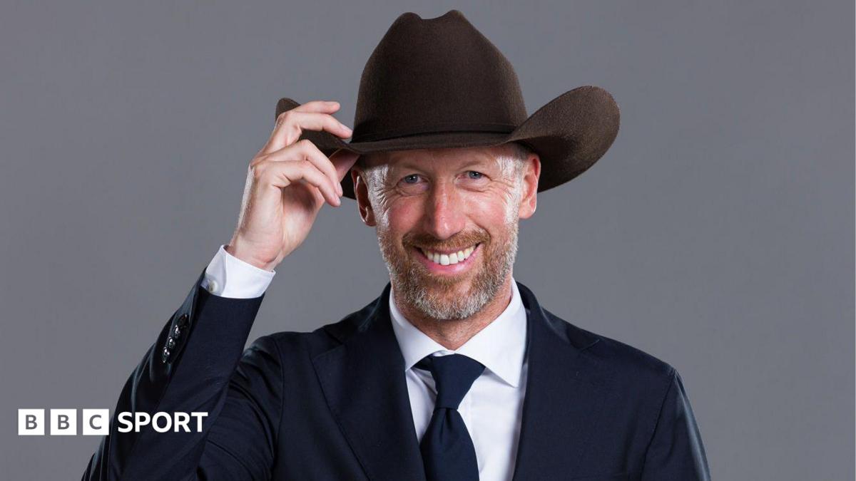 Graham Potter poses for the camera, wearing a suit while touching the brim of a cowboy hat