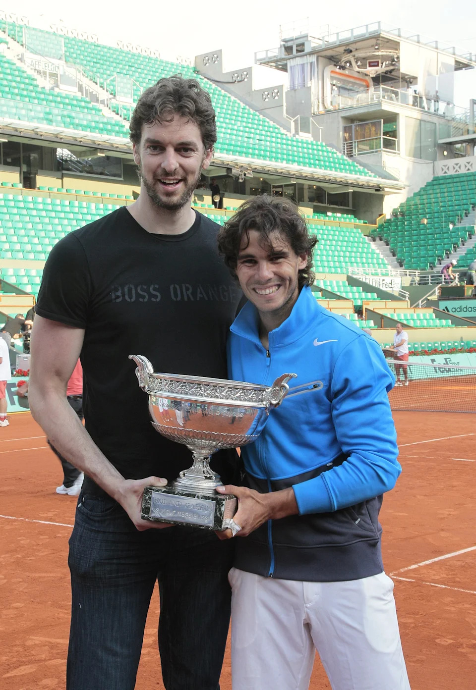 Two athletes holding a tennis trophy on a clay court, smiling, one in a casual t-shirt and jeans, the other in a sports jacket and shorts
