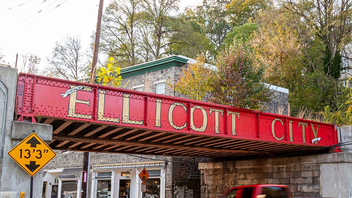 Welcome to Ellicott City sign on side of B&O railroad bridge with old houses and cars nearby