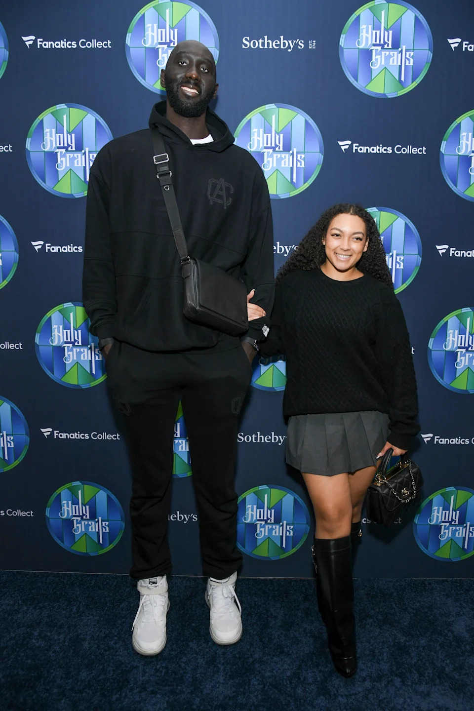 Two people stand together; one tall in a casual sweatshirt and sneakers, the other in a sweater and skirt with knee-high boots, at a sports event