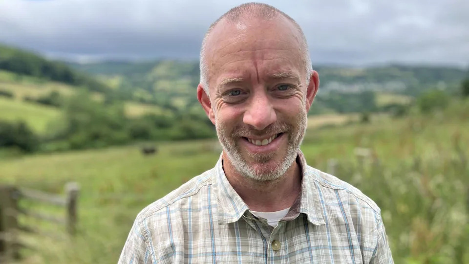 Man with thinning hair smiling at the camera. He has a moustache and beard and is wearing a check shirt with a white t-shirt under it. 