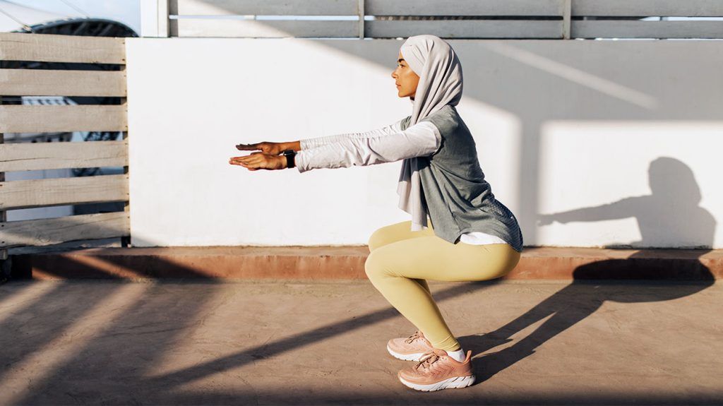 A person wearing athletic gear and a hijab does pilates outdoors in sunlight