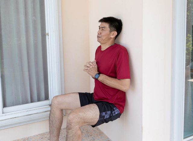 An adult Asian man performs a wall sit exercise against a wall at home during daytime. Suitable for concepts related to home workout, fitness routine, leg strength, and active lifestyle.