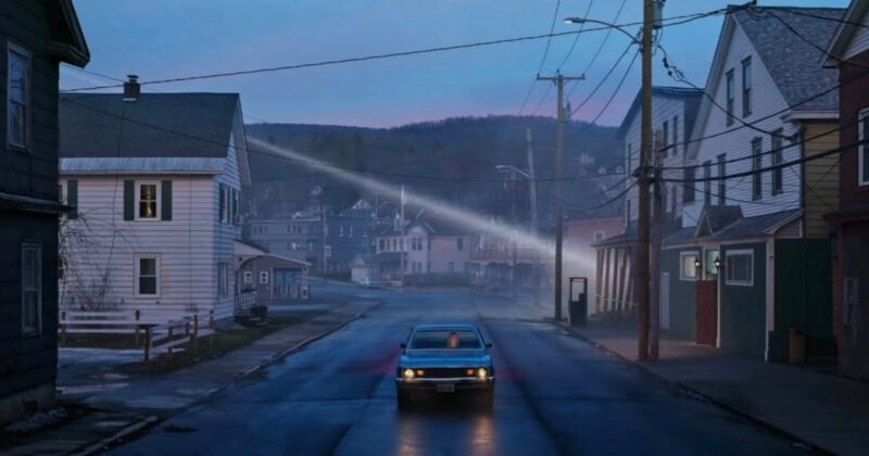 A blue car drives down a deserted street lined with houses at dusk; the sky is fading blue, power lines crisscross overhead, and distant hills and mist create a moody, quiet atmosphere.