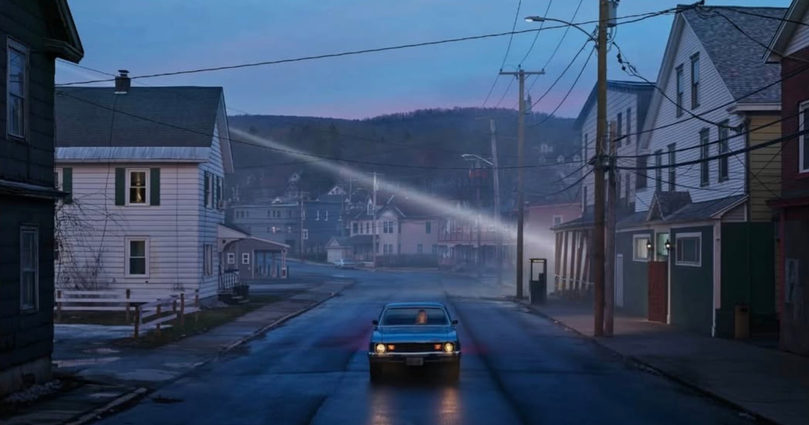 A blue car drives down a deserted street lined with houses at dusk; the sky is fading blue, power lines crisscross overhead, and distant hills and mist create a moody, quiet atmosphere.