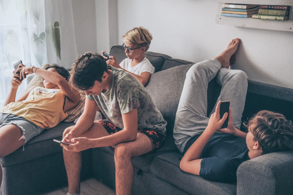 Four teenagers relaxing on a sofa while using their smartphones.