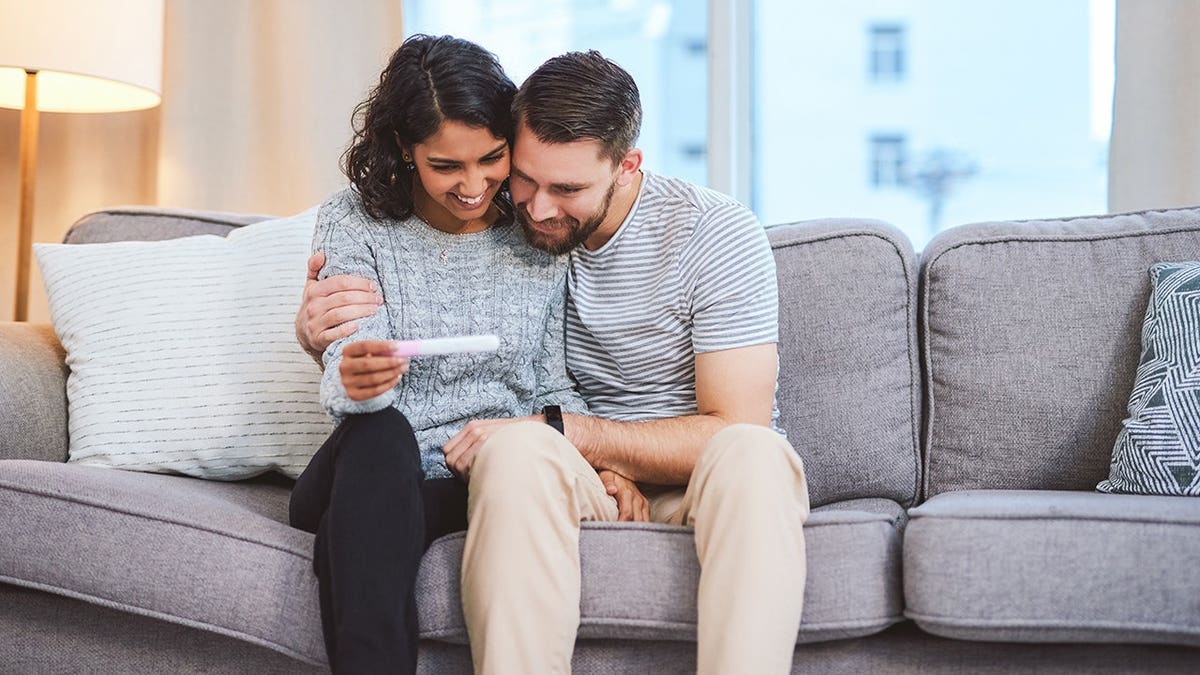 A happy young couple sitting on a sofa holding a positive pregnancy test.