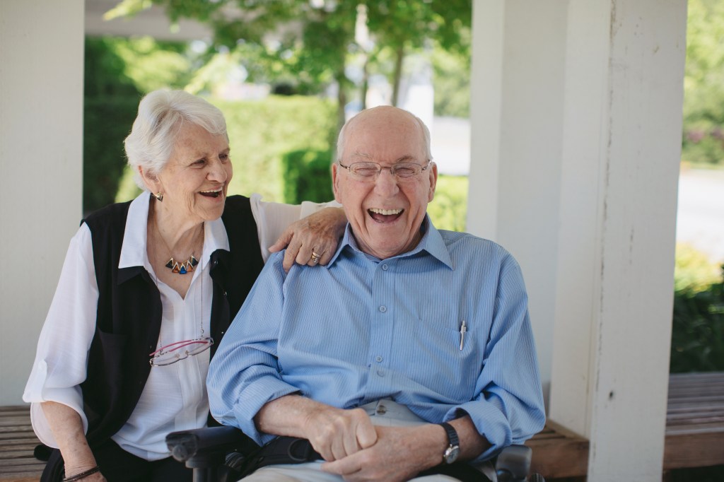 Happy elderly couple laughing, with the man in a wheelchair and the woman standing with her arm around him.