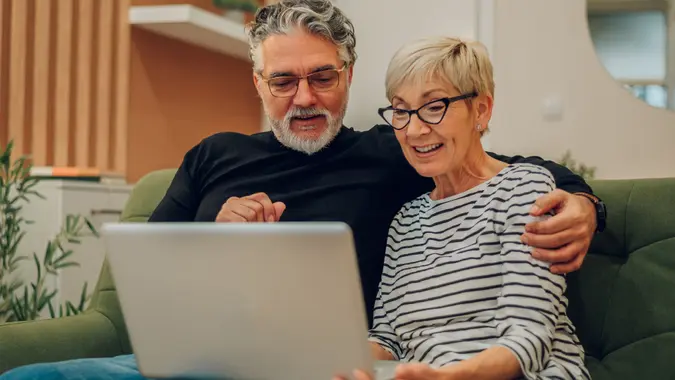 Senior couple leaning towards each other using laptop while sitting on a couch in their home stock photo