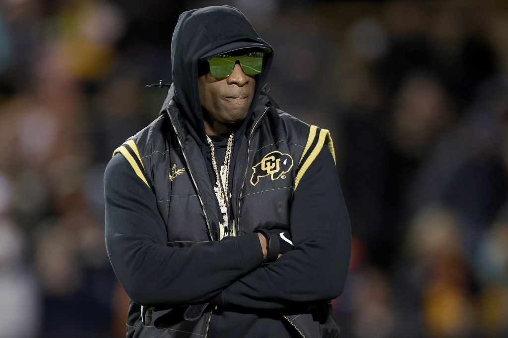 Deion Sanders, wearing a black hooded vest and green reflective sunglasses, stands on the field with his arms crossed.