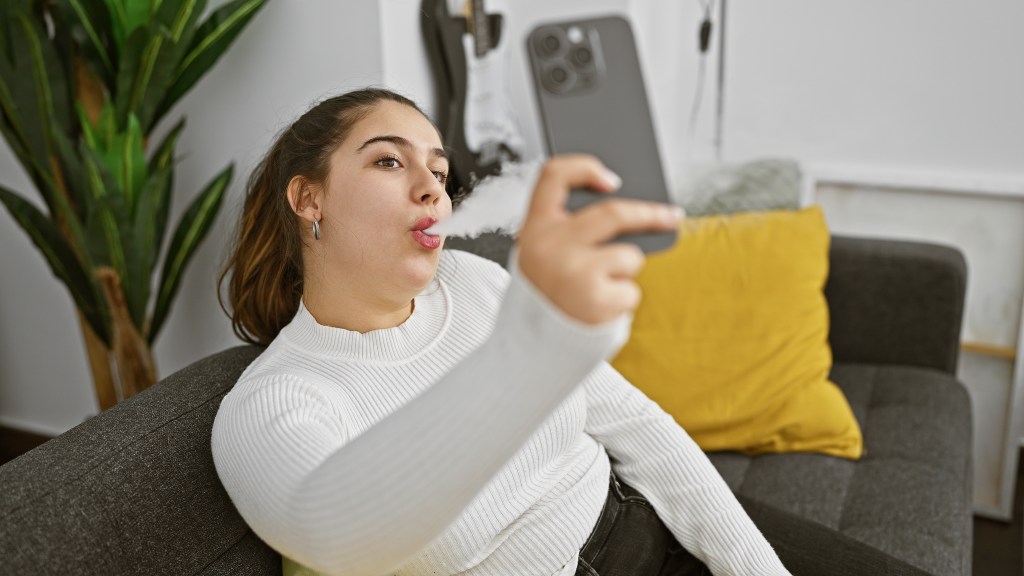 Hispanic woman vaping and taking a selfie on her smartphone in a cozy living room.