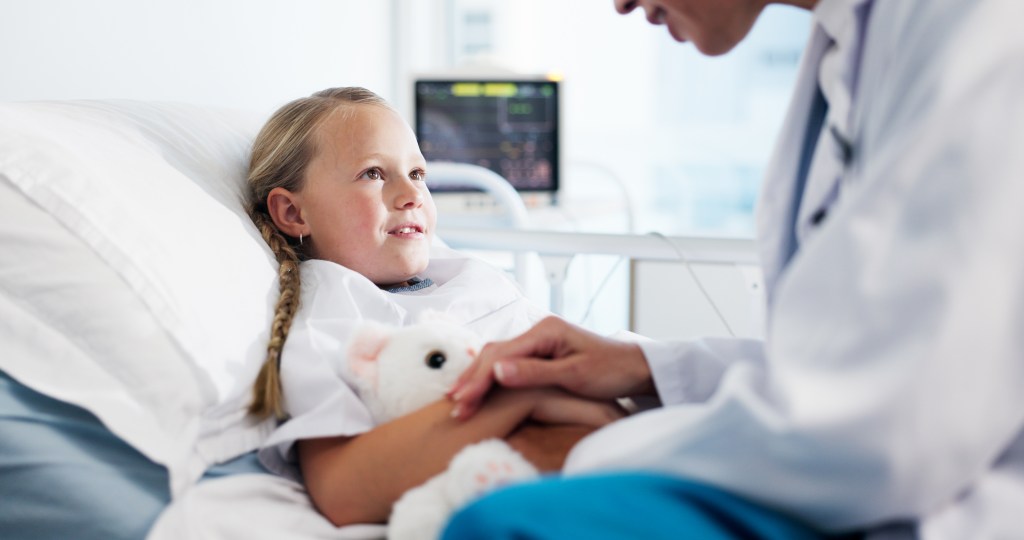 A doctor holding the hand of a child patient with a teddy bear in a hospital bed.