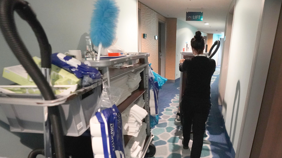 A woman is seen in a hotel hallway with a cleaning cart.