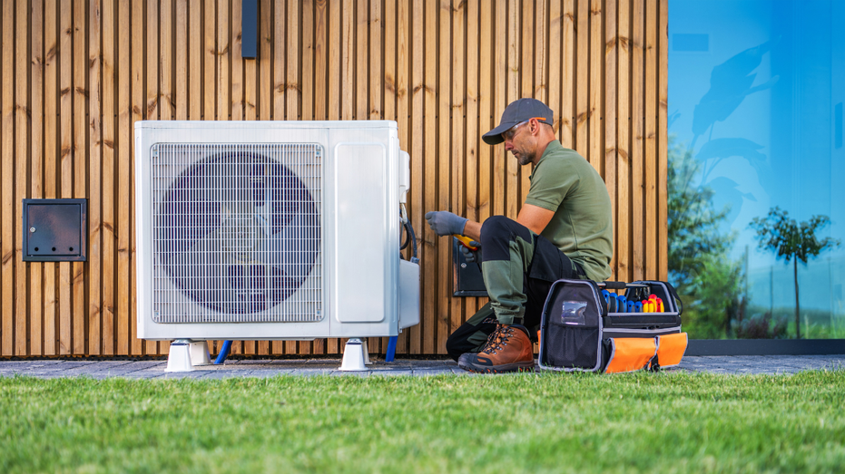 An HVAC technician is servicing an air conditioning unit located beside a modern home.