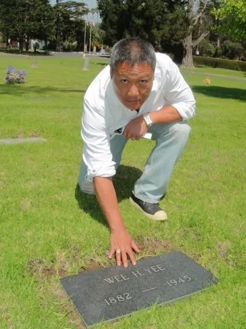 A man crouches on grass, touching a gravestone that reads "WEE H. YEE 1882 — 1945" in a cemetery.
