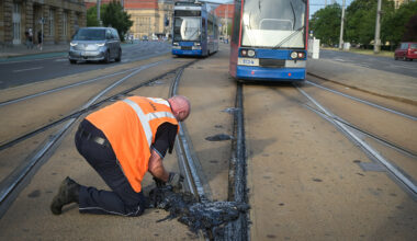 A worker in an orange safety vest examines heat-damaged tram tracks in Leipzig, where melted bitumen buckled under extreme temperatures as trams pass in the background.