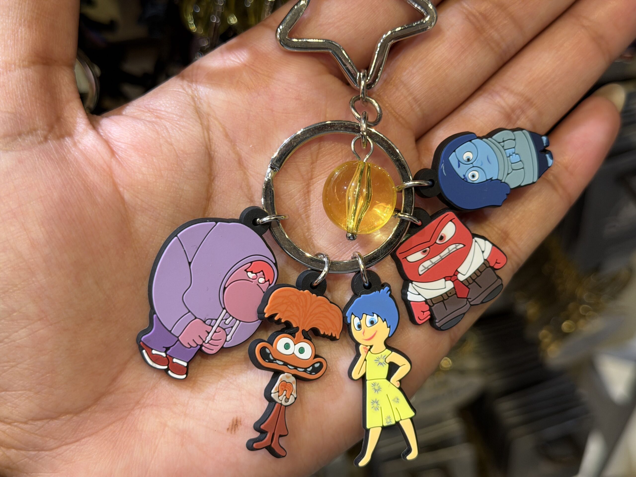 This keychain shows five characters from the Inside Out franchise. These figural characters are Embarrassment, Anxiety, Joy, Anger, and Sadness. A yellow, translucent ball, representing a joyful core memory, hangs in the center ring.