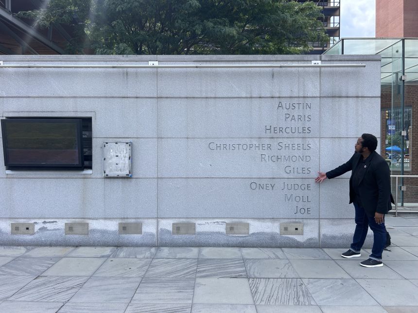Tour guide Mijuel Johnson points to the names of enslaved people etched into the President’s House exhibit near a removed panel.