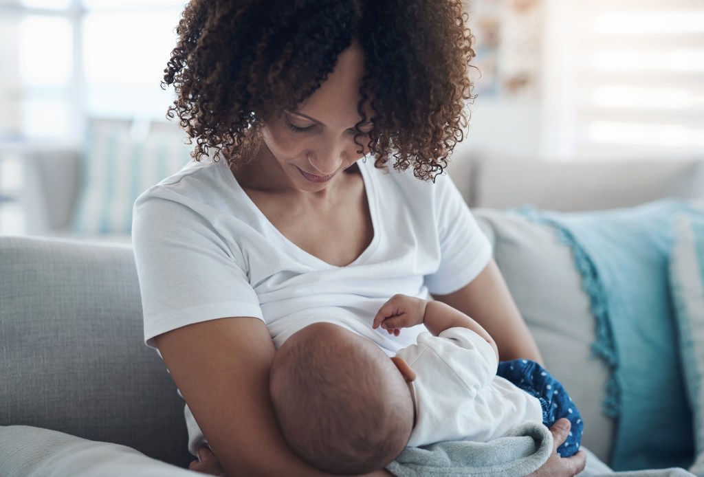 A mother with curly hair breastfeeds her baby on a couch.