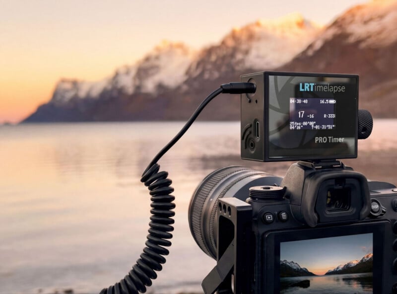 A camera with a time-lapse controller is set up by a calm lake at sunset, capturing snow-capped mountains and a colorful sky reflected in the water. The camera screen shows the scenic view being photographed.