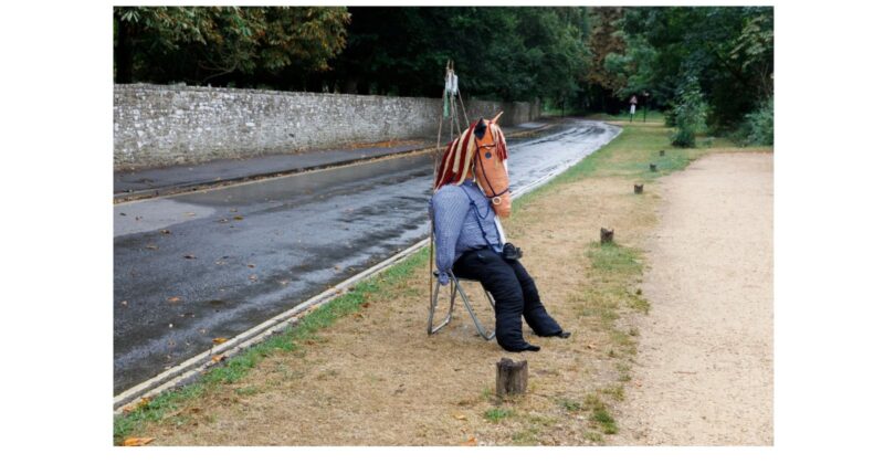A person wearing a horse mask, blue shirt, and black pants sits on a folding chair beside a wet road and a gravel path, with trees and a stone wall in the background.