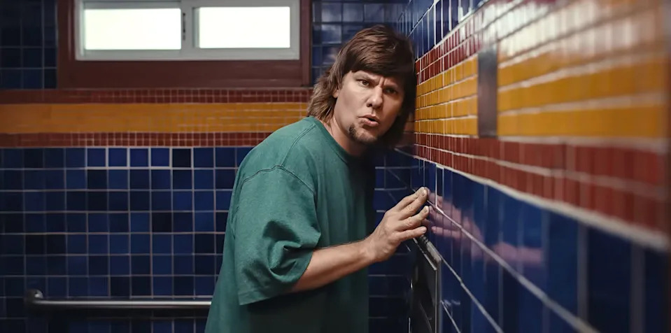 Theo Von leans against a tiled bathroom wall to listen in while pursing his lips in a scene from "Busboys."