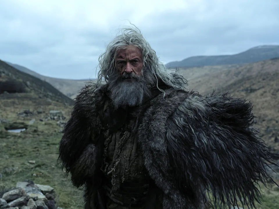 A gray-haired and heavily bearded Hugh Jackman wears a woolly coat while standing against a hilly backdrop.