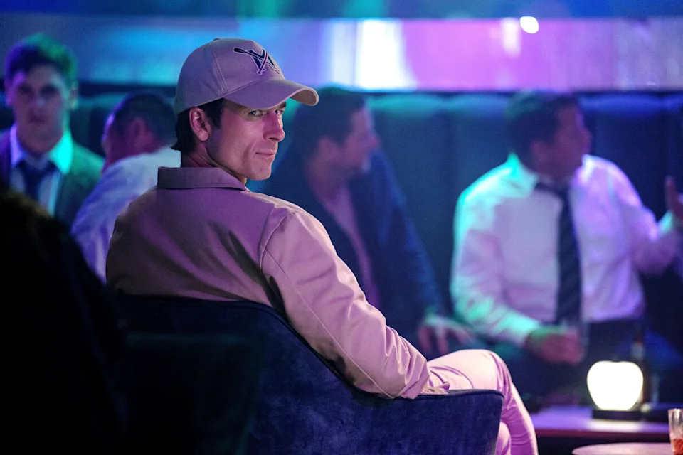 Glen Powell in a scene from "How to Make a Killing." He is sitting in a dark, crowded lounge.