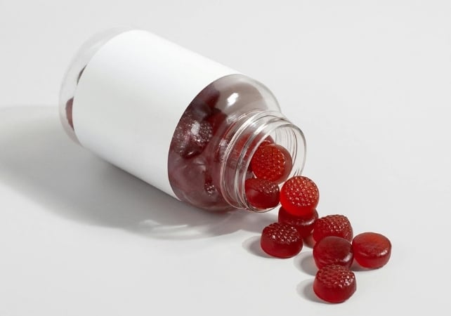 A white plastic supplement bottle lies on its side on a darker surface, spilling small red gummy supplements from the open neck.